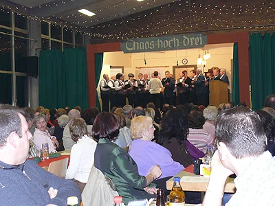 Männerchor singt auf einer Bühne im Saal, davor sitzt das Publikum
