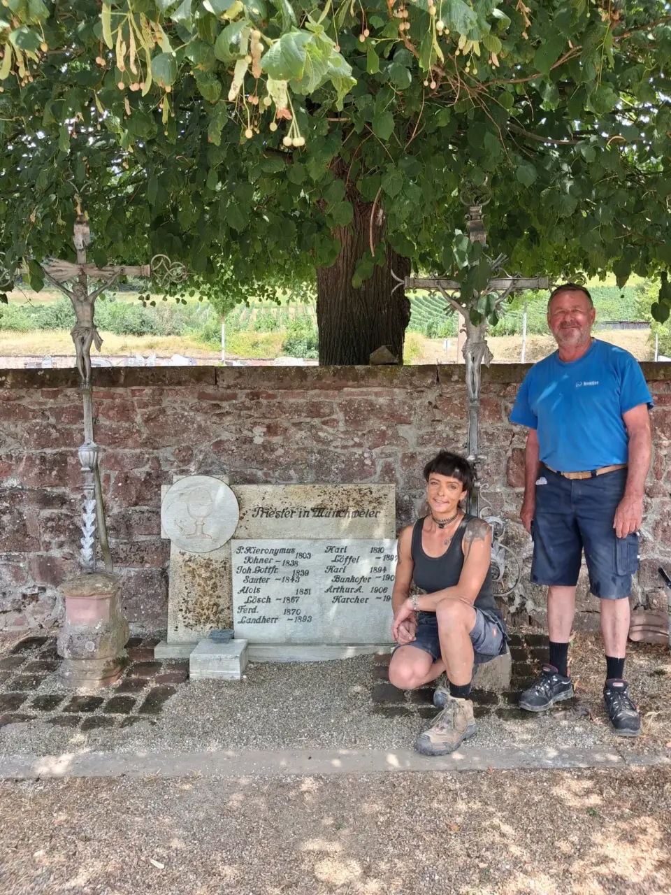 Zwei Personen vor einer Friedhofsmauer mit Kruzifix und einem restaurierten Grabstein
