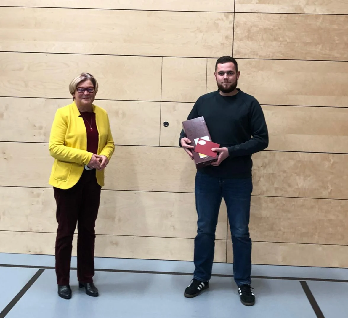 Frau in gelber Jacke und Mann mit einer Urkundenmappe vor einer Holzwand