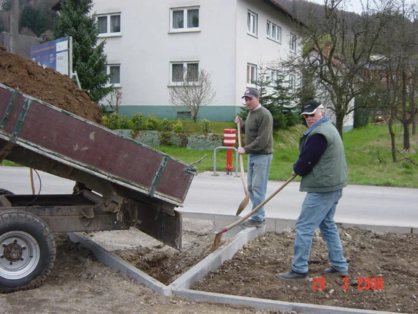 Zwei Männer schaufeln Erde neben einem Kipper-Anhänger