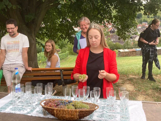 Frau in roter Jacke bei einer Weinprobe an einem Tisch mit Gläsern und Trauben, dahinter Zuschauende