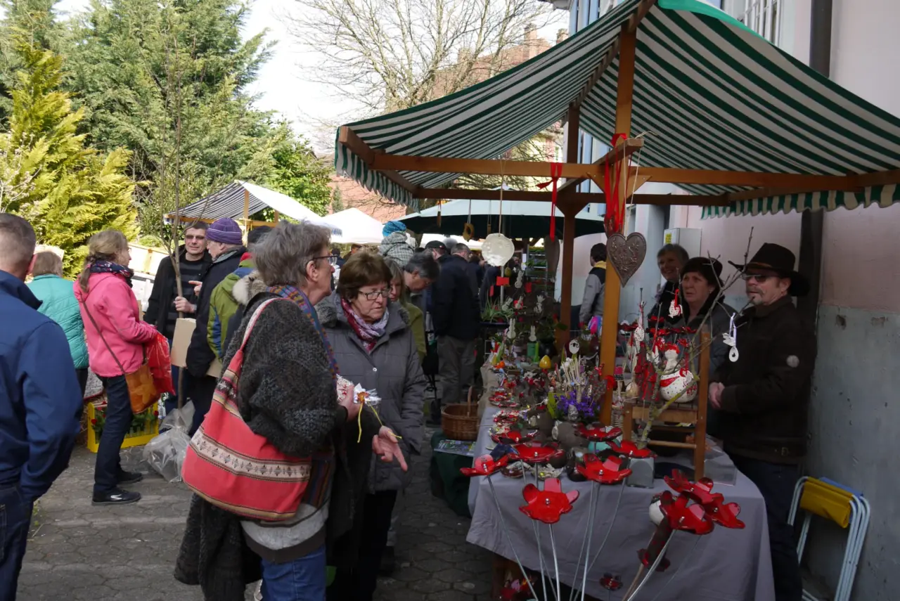 Marktstand mit roten Dekoblumen unter einer grün gestreiften Markise, davor Besucherinnen und Besucher