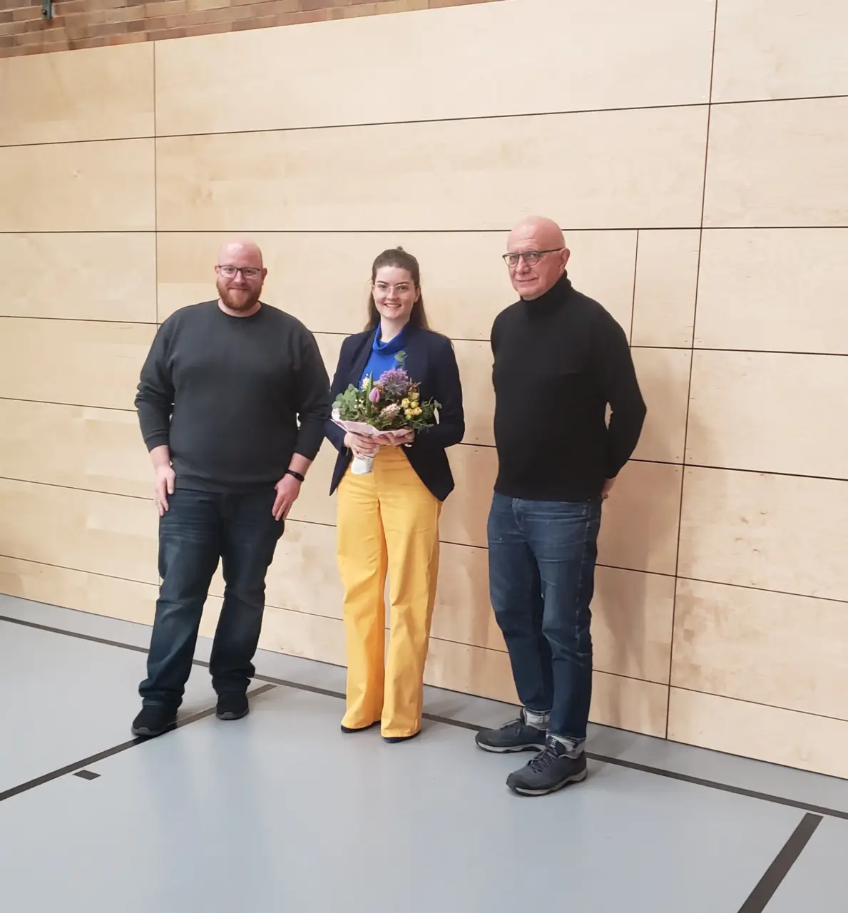 Drei Personen vor einer hellen Holzwand, die Frau in der Mitte hält einen Blumenstrauß