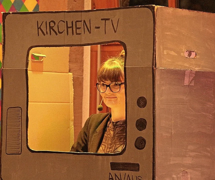 Frau schaut aus einem Pappfernseher mit der Aufschrift „Kirchen-TV“