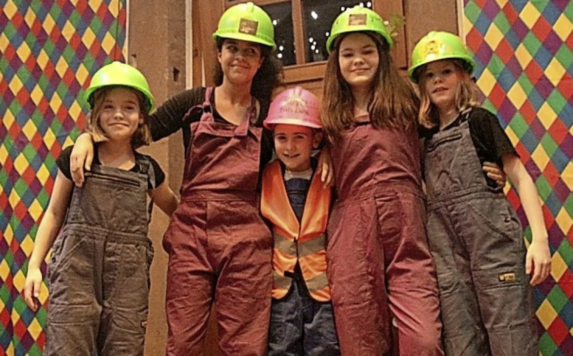 Kinder und eine Frau in Latzhosen mit grünen und rosa Bauhelmen