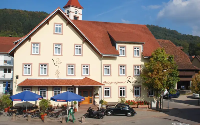 Gasthaus Rebstock: großes Gebäude mit Walmdach und Dachreiter, davor eine Terrasse mit Sonnenschirmen