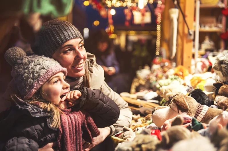Frau und Kind mit Wollmützen betrachten staunend Waren auf einem Weihnachtsmarkt