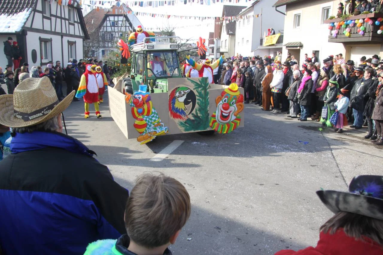 Fasentsumzug mit Festwagen auf einer Dorfstraße, Zuschauer am Straßenrand